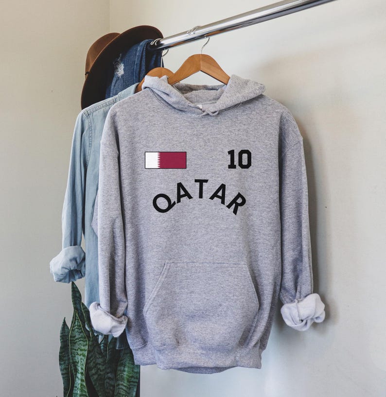 K&ouml;nnte beinhalten: Ein grauer Kapuzenpullover mit dem Wort "QATAR" in schwarzem, gebogenem Text. &Uuml;ber dem Text befindet sich ein weinrotes und wei&szlig;es Rechteck und die Zahl "10" in Schwarz. Der Kapuzenpullover h&auml;ngt an einem Holzb&uuml;gel.