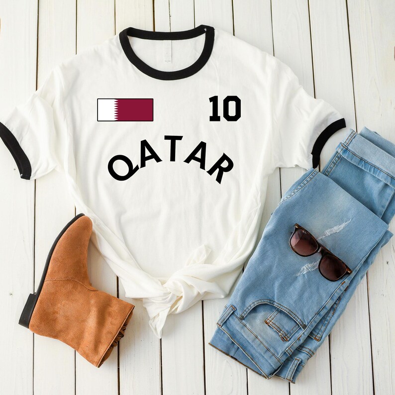 K&ouml;nnte beinhalten: Wei&szlig;es T-Shirt mit schwarzem Besatz und dem Wort "QATAR" in gro&szlig;en schwarzen Buchstaben. Die Nummer 10 und eine Katar-Flagge sind ebenfalls aufgedruckt. Hellblaue Jeans, braune Stiefel und Sonnenbrille sind ebenfalls im Bild.