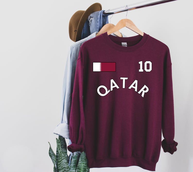May include: A maroon sweatshirt with the word "QATAR" in white arched across the front. Above the word is a rectangular graphic of the Qatar flag and the number "10". The sweatshirt is hanging on a wooden hanger.