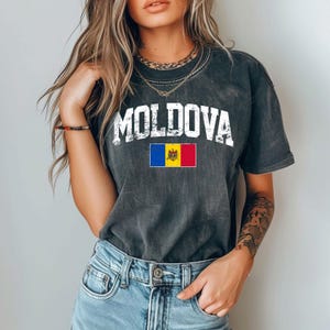 Könnte beinhalten: Dunkelgraues T-Shirt mit dem Wort "MOLDOVA" in weißen Blockbuchstaben über der moldauischen Flagge. Die Flagge hat blaue, gelbe und rote vertikale Streifen mit einem Wappen in der Mitte. Das Shirt wird mit blauen Jeans getragen.