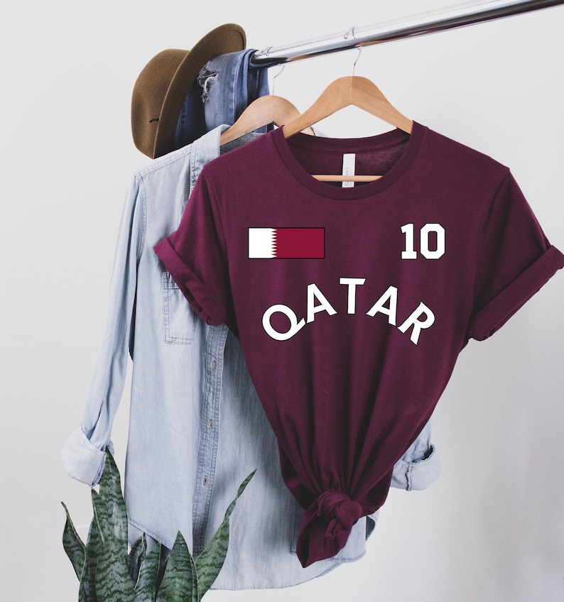 May include: A maroon t-shirt with the word "QATAR" in white, arched across the front. Above the word is a rectangular graphic of the Qatar flag and the number "10". The shirt is hanging on a wooden hanger.