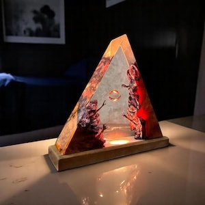 May include: A unique, triangular resin lamp with a wooden base. Inside, a gold ring floats between two reddish-brown, textured sculptures. The lamp emits a warm, inviting glow, creating a captivating display.