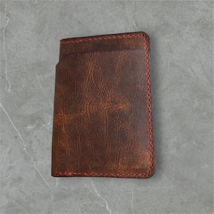 May include: A brown leather card holder with red stitching. The wallet has a distressed appearance and a simple design. The card holder is rectangular and appears to have multiple card slots.
