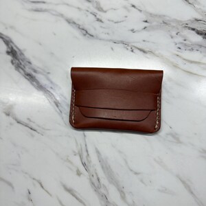 May include: A brown leather card holder with a flap closure. The card holder has three card slots and white stitching around the edges. The leather has a smooth texture.