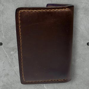 May include: A dark brown leather wallet with visible stitching in a lighter brown color. The wallet is rectangular and appears to be folded. The edges are rounded. The background is a mottled gray.