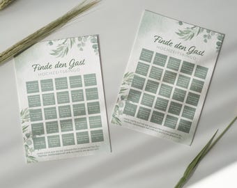 以下が含まれることがあります： 「Finde den Gast HOCHZEITSBINGO」の文字が入ったウェディングビンゴカードが2枚。カードにはテキストが入った正方形のグリッドがあり、水彩画風の緑の葉で装飾されています。