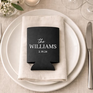 Könnte beinhalten: Ein schwarzer Getränkedosenkühler mit dem Text "the WILLIAMS" und dem Datum "2.18.24" in Weiß. Der Kühler liegt auf einem weißen Teller mit einer gefalteten beigen Serviette. Eine Gabel und ein Messer liegen auf dem Teller.