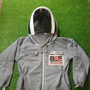 May include: A gray beekeeping suit with a white hood and a clear face shield. The suit has a checkered pattern, multiple pockets, and a zipper down the front. The "BEE STUFF ENTERPRISES" logo and an American flag patch are visible.