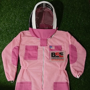 May include: Pink beekeeping suit with a white mesh hood. The suit has a zipper closure, pockets, and elbow patches. The text "BEE STUFF ENTERPRISES" is on the chest, with an American flag patch.