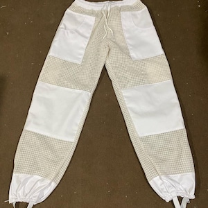 Puede incluir: Pantalones de apicultura blancos y beige. Los pantalones tienen bolsillos y parches blancos en las piernas, con tejido de malla beige en el medio. Cuentan con una cintura elástica con cordón y estribos en los tobillos.