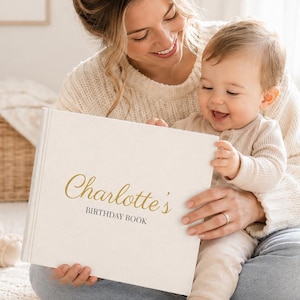 May include: A white hardcover book with gold foil lettering on the front cover that reads Charlotte's BIRTHDAY BOOK. The book has a clean, textured surface and is held open to display the title on the front cover in a clear, legible font.