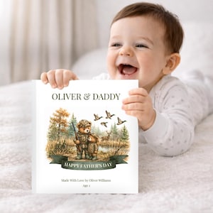 May include: A white card with the text "OLIVER & DADDY" and "HAPPY FATHER'S DAY" is held by a baby. The card features an illustration of two teddy bears in hunting attire, with ducks and deer in the background. The card also includes the text "Made With Love by Oliver Williams" and "Age 1".