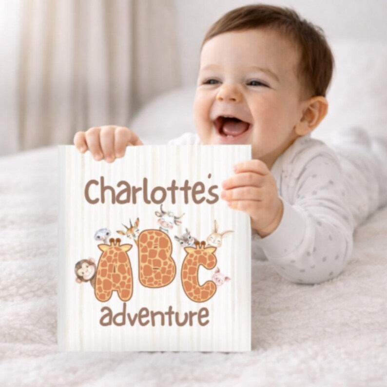 May include: A happy baby holds a children's book. The book cover reads "Charlotte's ABC adventure" with animal illustrations around the letters. The letters A, B, and C are designed with giraffe patterns. The background has a light, striped pattern.