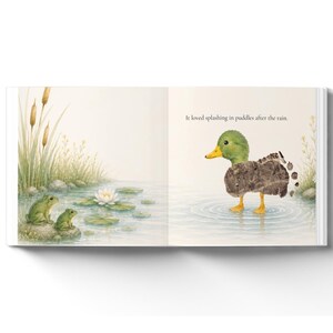 May include: An open children's book page featuring a watercolor illustration of two green frogs on a rock near a white water lily and a duckling covered in mud standing in water. The text reads: It loved splashing in puddles after the rain.