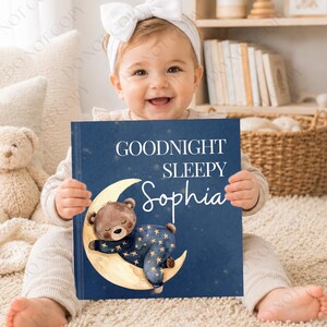 Może przedstawiać: Spersonalizowana książka dla dzieci z granatową okładką z napisem "Goodnight Sleepy Sophia". Ilustracja na okładce przedstawia śpiącego misia na księżycu w kształcie półksiężyca. Książkę trzyma dziecko.