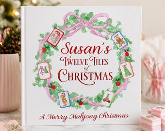 May include: A white square book cover featuring a watercolor green wreath with a pink ribbon. The text reads Susan's TWELVE TILES of CHRISTMAS and A Merry Mahjong Christmas. Mahjong tiles with holiday illustrations are placed around the wreath.