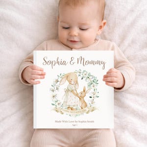 May include: A baby holds a book with the text "Sophia & Mommy" above a watercolor illustration of two bunnies inside a floral wreath. The book also includes the text "Happy Mother's Day", "Made With Love by Sophia Smith", and "Age 1".