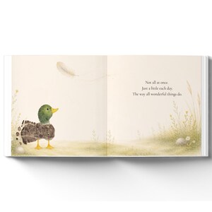 May include: An open book page featuring a watercolor illustration of a mallard duck with a green head and yellow feet standing in a grassy field. A single feather floats above. The text reads: Not all at once. Just a little each day. The way all wonderful things do.