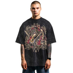 Puede incluir: Camiseta gris oscuro de gran tamaño con un gráfico detallado de una criatura estilizada con acentos rojos y dorados. El diseño está centrado en la parte delantera, con intrincados patrones que rodean la figura central. La camiseta tiene mangas cortas.