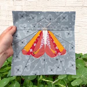 May include: A square fabric block featuring a colorful butterfly design. The butterfly wings are made of orange, red, and white patterned fabric, set against a gray background with a white floral pattern. The block is held by a hand.