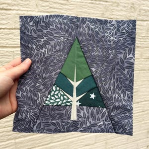 Op de afbeelding: Vierkant stoffen stuk met een gestileerd boomontwerp. De boom is samengesteld uit verschillende tinten groen en wit, met een witte ster. De achtergrond is een grijze stof met een bladerenpatroon.