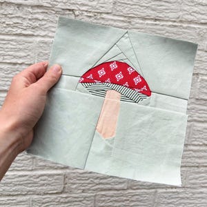 May include: A handmade fabric square featuring a mushroom design. The mushroom cap is red with a white diamond pattern, the stem is beige, and the base fabric is light green. The design is likely part of a larger quilt or textile project.
