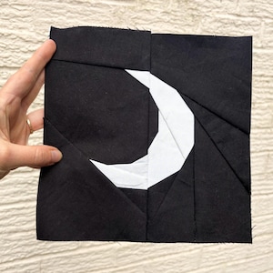 May include: A black and white fabric square featuring a crescent moon design. The moon is white, set against a black background. The fabric is held, highlighting the geometric pattern and contrasting colours.