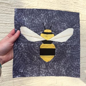 May include: A square fabric block featuring a stylized bee design. The bee has a yellow and black body, white wings, and is set against a blue background with a leaf pattern. The block is held by a hand.