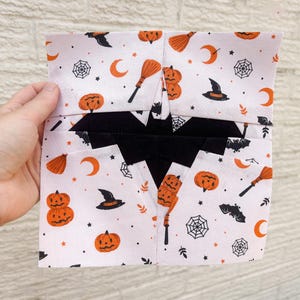 May include: A folded Halloween-themed fabric featuring a white backdrop with orange and black illustrations. The design includes pumpkins, witch hats, bats, spiderwebs, brooms, and crescent moons. The fabric is held by a hand.
