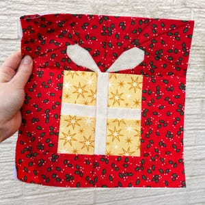 May include: A handmade Christmas gift block. The square block features a red background with a holly berry pattern. A yellow gift box with gold star accents is centered, with white ribbon and bow details.