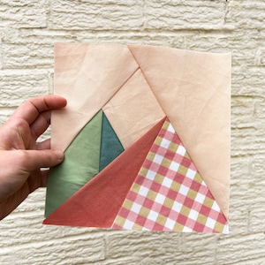 May include: A geometric fabric block with a patchwork design. The block features a light peach background with triangular sections of green, teal, red, and a red and white gingham pattern. The block is held by a hand.