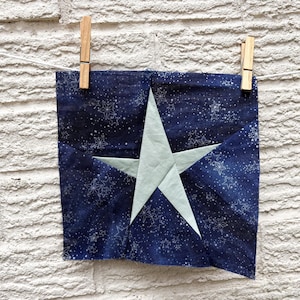 May include: A square fabric piece with a navy blue background and a light blue star design. The fabric is decorated with white snowflake patterns. The piece is held by two wooden clothespins on a white string.