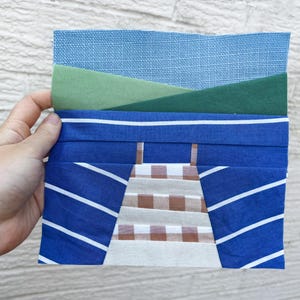 May include: A fabric art piece featuring layered textiles. The top layer is a textured light blue, followed by green and dark green sections. Below, a blue fabric with white diagonal stripes and a central design of brown and white checkered fabric.