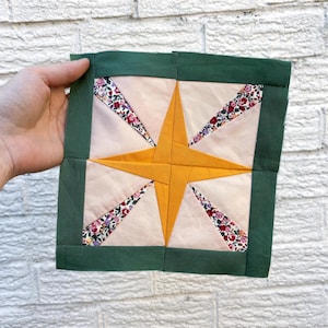 May include: A square patchwork quilt block with a star design. The star is yellow, set against a light pink background, with floral print accents. The edges are framed with a dark green border. The block is held against a white brick wall.