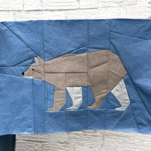 May include: A blue and tan fabric art piece featuring a geometric bear design. The bear is depicted in shades of tan and gray, set against a blue background. The artwork is held up by hands.