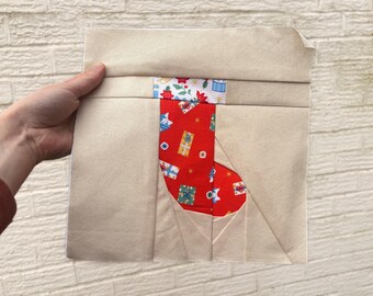 Op de afbeelding: Een handgemaakt kerstsokblok met een rood sokontwerp. De sok is versierd met geschenkdozen en sterren. Het blok is gemaakt van beige stof en wordt door een hand vastgehouden.