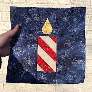 May include: A quilted Christmas candle design on a dark blue background with white snowflakes. The candle has a red and white striped body with green holly accents and a yellow flame.