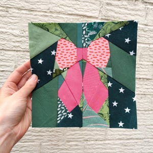 Peut inclure: Un bloc de courtepointe carré fait à la main représentant un nœud rose. Le bloc est composé de divers tissus verts, notamment des teintes unies, des imprimés floraux et un tissu bleu marine avec des étoiles blanches. Le nœud a un motif à pois.