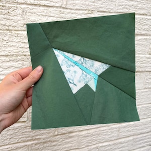 Puede incluir: Una pieza de arte textil geométrica con un diseño de montaña. La obra de arte es principalmente verde oscuro, con una sección central de tela blanca estampada con elementos azules y turquesas. La pieza se sostiene contra una pared de ladrillos blancos.