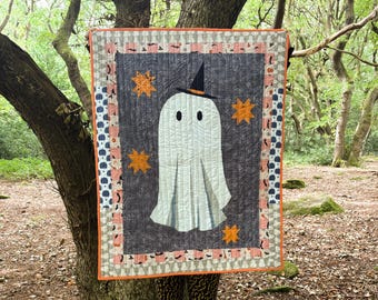 Könnte beinhalten: Ein Halloween-Quilt mit einem weißen Geist, der einen schwarzen Hexenhut trägt, vor einem dunkelgrauen Hintergrund mit orangefarbenen Sternakzenten. Der Quilt hat einen gemusterten Rand mit orangefarbenem Besatz, ideal für die saisonale Dekoration.