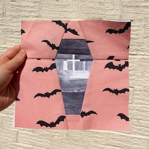 May include: A folded paper craft with a Halloween theme. The pink paper is decorated with black bat silhouettes. The center reveals a coffin shape with a cross, painted in shades of gray and white.