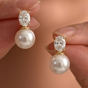 May include: A pair of earrings with a large, white pearl and a sparkling, oval-cut cubic zirconia. The earrings have a gold-coloured setting. The jewellery is held up by a person.