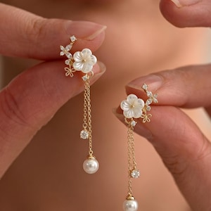 Könnte beinhalten: Goldfarbene Ohrringe mit floralem Design, weißen Blütenblättern und funkelnden Akzenten. Zarte Ketten mit einer Perle am Ende. Die Ohrringe werden von Händen gehalten.