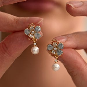 May include: A pair of gold-coloured flower earrings. Each earring features a light blue flower design with clear crystal accents and a dangling white pearl. The earrings are held up by a person's hands.