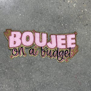 May include: Pink and gold glitter text that reads "BOUJEE on a budget". The text is outlined in black and has a gold lightning bolt design on the sides. The background is a grey, textured surface.