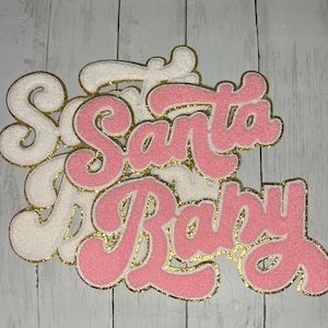 May include: Fuzzy white and pink felt letters spelling "Santa Baby" with gold glitter edging. The letters are layered, with a textured appearance. The background is a light wood grain pattern.