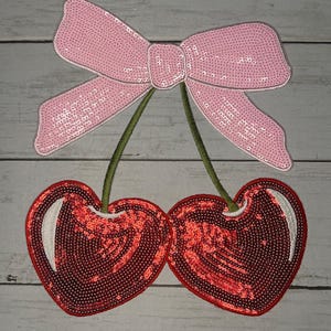 Può includere: Una toppa decorativa con un paio di ciliegie rosse con paillettes con steli verdi, sormontate da un fiocco rosa con paillettes. Le ciliegie hanno un riflesso bianco. La toppa è progettata per l'abbellimento.