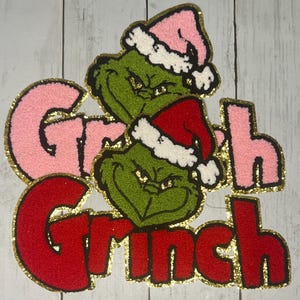 Peut inclure: Un motif festif sur le thème du Grinch avec le mot "Grinch" en rouge et rose, souligné de paillettes dorées. Le visage du Grinch est vert avec un bonnet de Père Noël, parfait pour les créations de vacances.