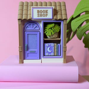 May include: A miniature book shop model with a purple door and window frames. The sign above the shop reads "BOOK SHOP". Inside the shop, there are miniature books and a potted plant. The model is set on a stack of pink books.