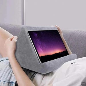 May include: A grey tablet pillow supporting a tablet showing a purple and orange sunset. The pillow is made of a soft material and is designed to support a tablet. The tablet is held in place by the pillow's design, ideal for comfortable viewing.